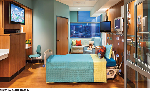 A colorful children's hospital room.  The walls and bed are teal with yellow accents.  There is extensive wood paneling on the walls and a teddy bear sits on the chair.
