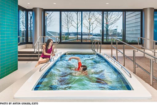 A physical therapist sits next to a therapy pool as a patient performs physical therapy exercises in the water
