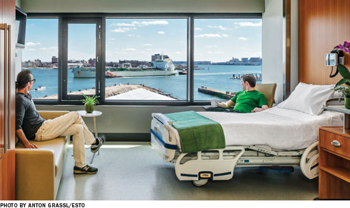 Two people sit in chairs in a hospital room and look out the window at a boat on the water