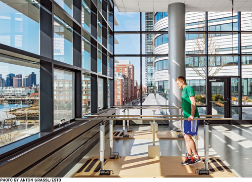 A patient performs physical therapy walking exercises on parallel bars in a sunny room with floor to ceiling windows