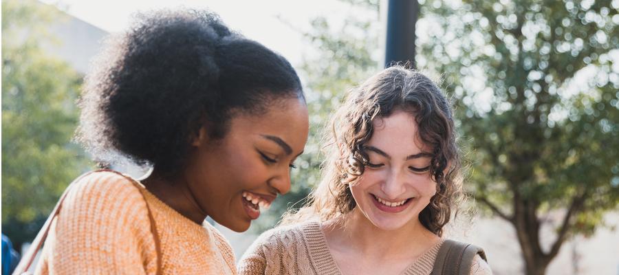 A Black teenage girl and a white teenage girl laugh while looking at a phone.