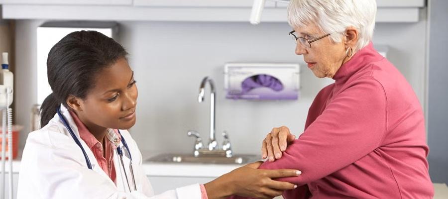 A female physician examines the elbow of a geriatric female patient in an examination room.